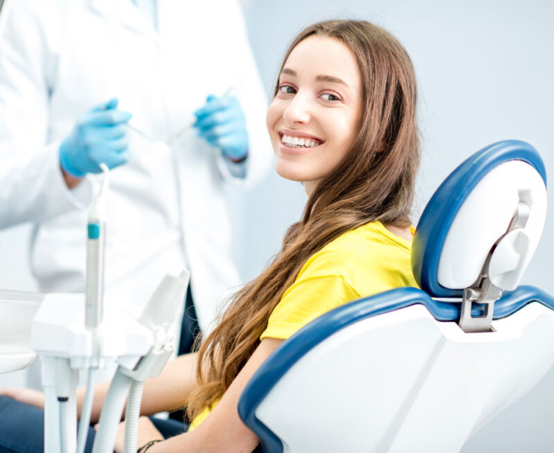 Brunette young woman in a yellow shirt smiles while sitting in a dental chair after soft tissue laser treated her gum disease