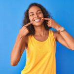Brown woman in a yellow tank top points to her smile after professional teeth whitening on a blue background
