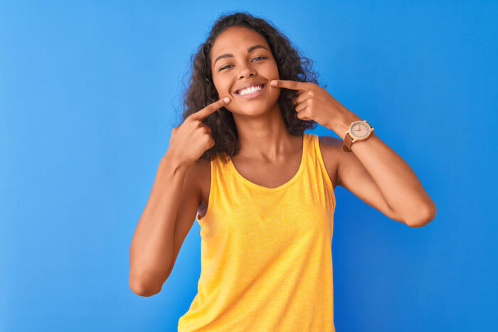 Brown woman in a yellow tank top points to her smile after professional teeth whitening on a blue background